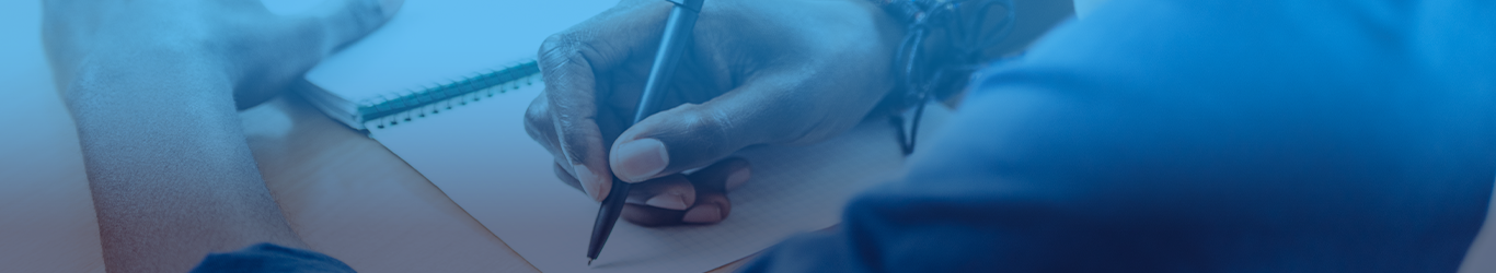 Image shows a man writing in a notepad on a desk.