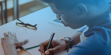 Image shows a man writing in a notepad on a desk.